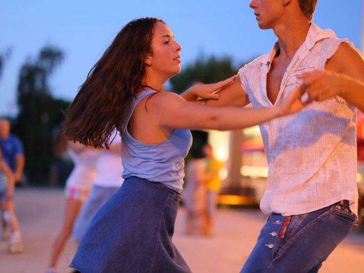 A young couple dances salsa outdoors at dusk, the leader guiding his partner across