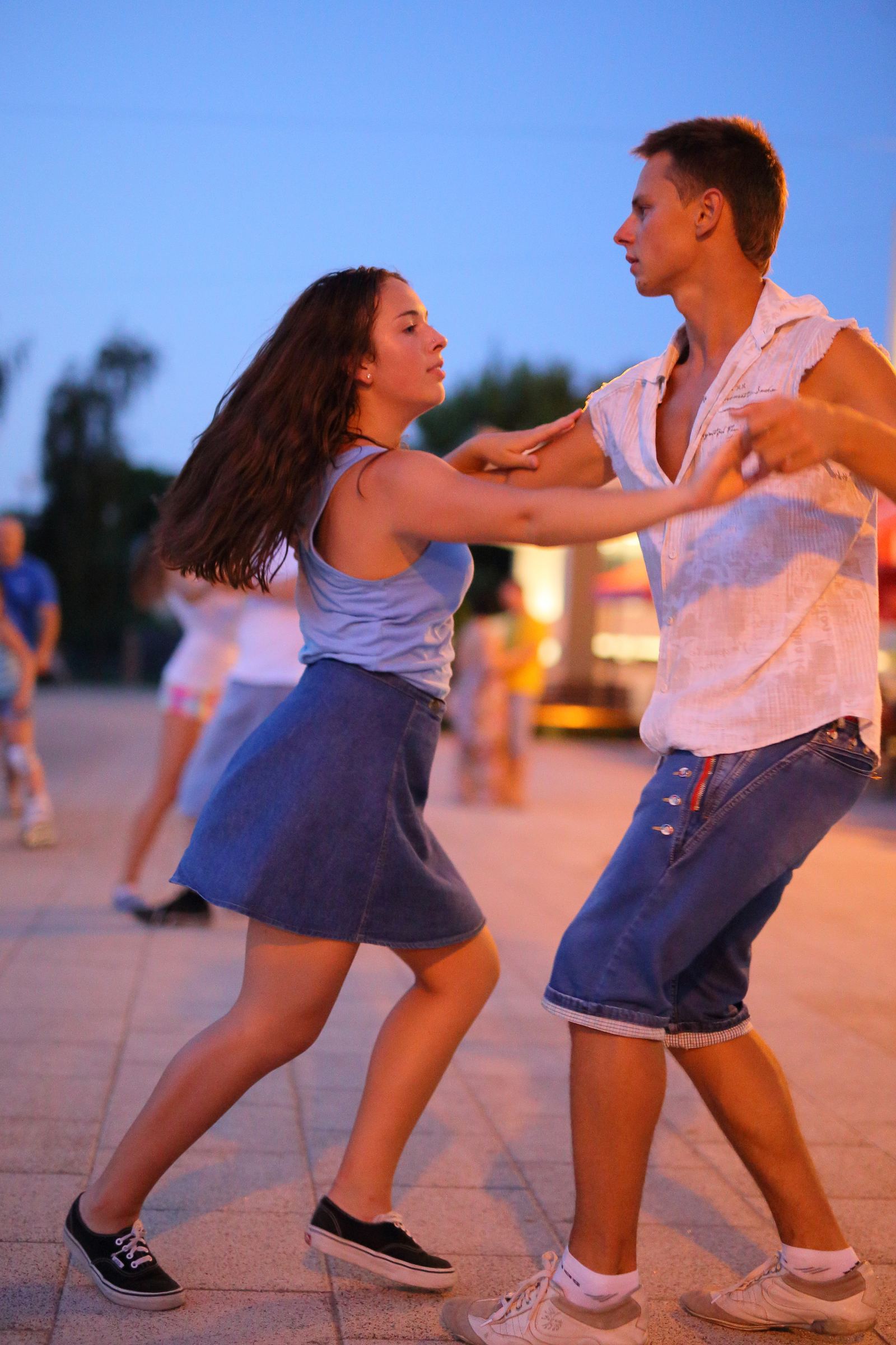 A young couple dances salsa outdoors at dusk, the leader guiding his partner across