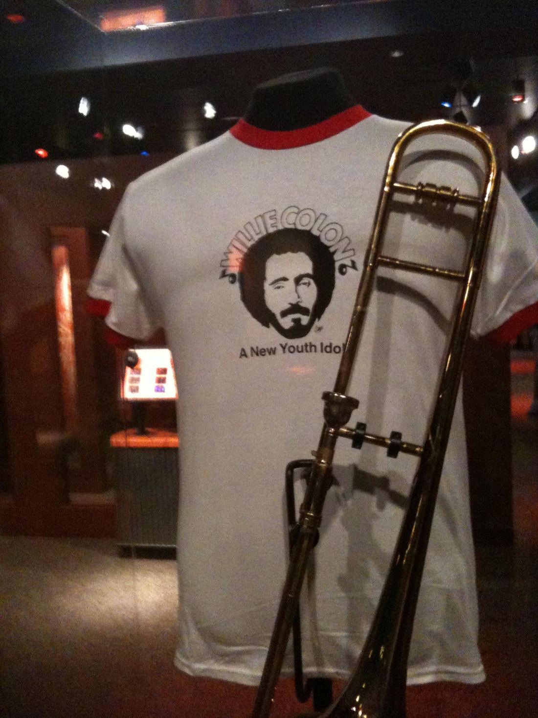 A museum display with a Willie Colón T-shirt and a trombone